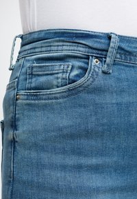 Gros plan de la zone de poche d'un jean en denim bleu avec des coutures visibles, des rivets, une boucle de ceinture et une partie d'une chemise blanche au-dessus de la taille.