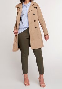 Beige trench coat s dvojitým zapínáním, límcem a dlouhými rukávy, nošený přes modro-bíle pruhovanou košili a olivově zelené kalhoty.