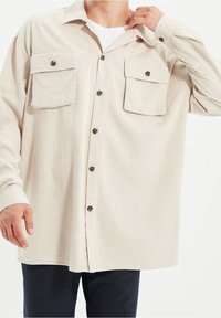 Lichtbeige, langemouw shirt gemaakt van zachte stof. Bevat twee voorkruiszakken, een knoopsluiting en donkere knopen als accent.