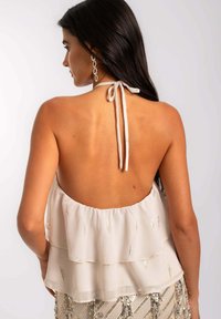 Vue arrière d'un haut halter beige avec deux couches transparentes, présentant des accents métalliques et un lien au cou, associé à une jupe pailletée.