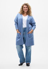Cardigan azzurro chiaro in maglia morbida, aperto davanti, con maniche lunghe e due tasche frontali, abbinato a jeans larghi e scarpe nere.