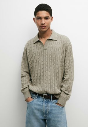 Junger Mann mit kurzen lockigen Haaren, der einen grauen Zopfpullover und hellblaue Jeans trägt, steht mit den Händen in den Taschen vor einem weißen Hintergrund.
