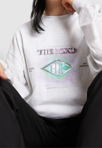 Ljust grå sweatshirt med en grafisk design som har texten "THE MIND" och en diamantlogotyp. Långa ärmar, avslappnad passform, mjukt tyg.