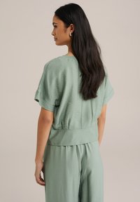 Top verde chiaro a maniche corte realizzato in tessuto testurizzato. Presenta una vestibilità comoda e un elastico in vita. Design semplice e minimalista.
