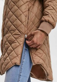 Quilted beige jacket with a zip closure, featuring ribbed cuffs and an asymmetrical hem, showing a hand adjusting the zipper.