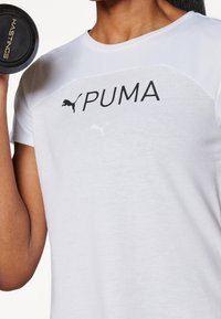 Camiseta deportiva blanca con un logo negro de PUMA, con acentos de malla en los hombros y las mangas cortas.