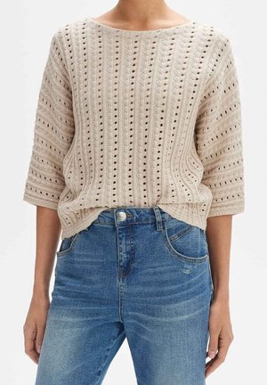 Pull beige en tricot à manches mi-longues avec des motifs de trous verticaux, rentré dans un jean en denim bleu délavé à taille haute.
