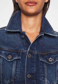 Mörkblå denimjacka med klassisk krage, två bröstfickor, silverfärgade metallknappar och kontrasterande sömmar.