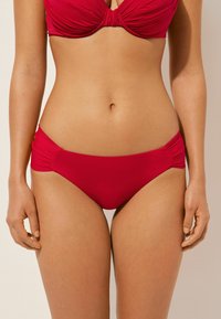 Ensemble bikini rouge avec un design froncé sur les côtés. Le matériau est lisse et extensible, avec un détail noué au centre du haut.