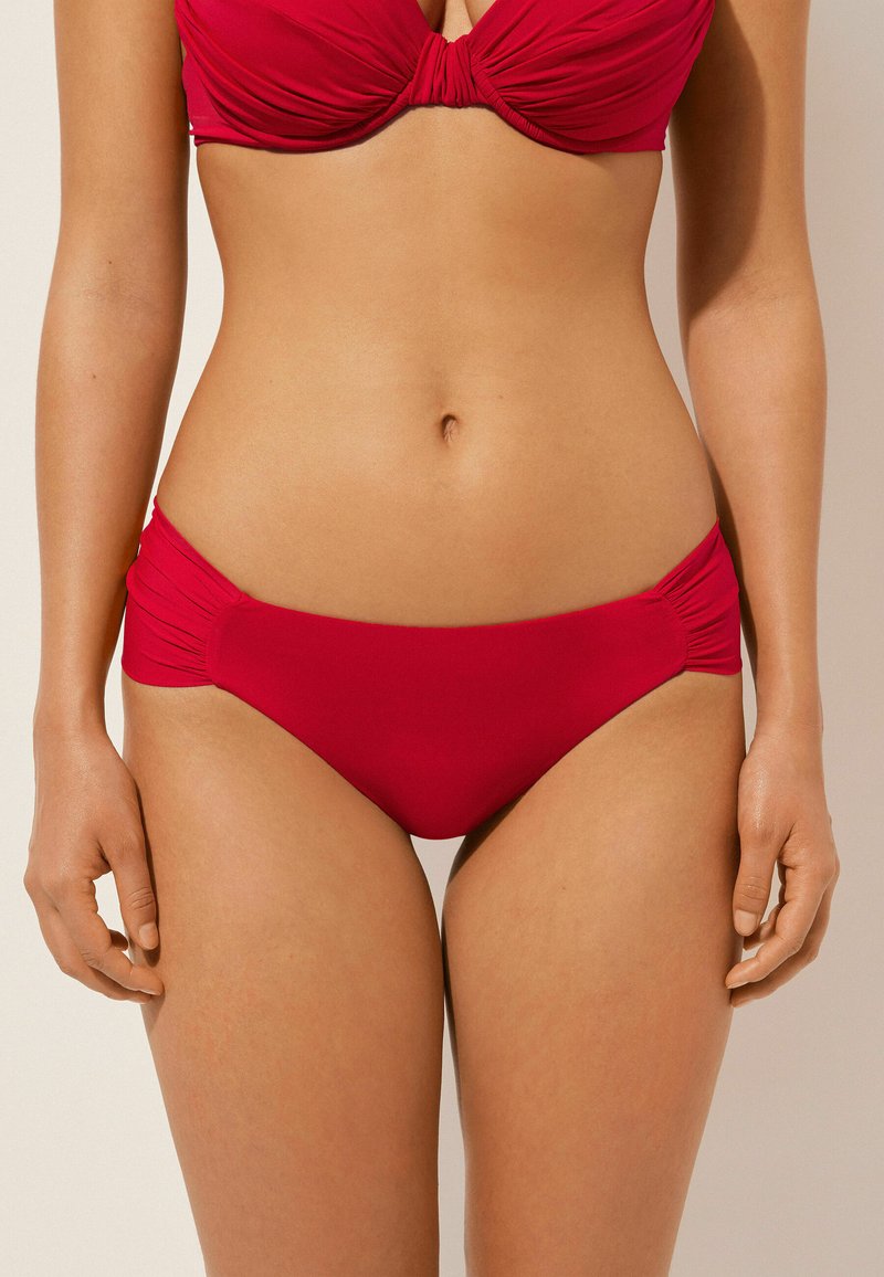 Ensemble bikini rouge avec un design froncé sur les côtés. Le matériau est lisse et extensible, avec un détail noué au centre du haut.