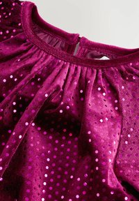 Fuchsiaklänning med en texturerad tyg, försedd med glittriga paljetter och en rynkad halsringning, som visar en nyckelhålsöppning bak.