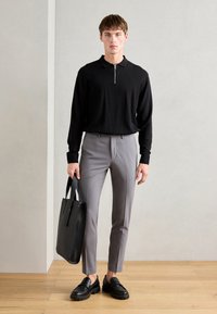 Black long-sleeve polo with a zip neck, paired with slim-fit gray trousers and black loafers; holding a black tote bag.