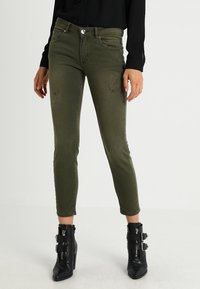Jeans ajustados en verde oliva con sutiles detalles desgastados, combinados con botas negras hasta el tobillo con hebillas y un acabado texturizado.