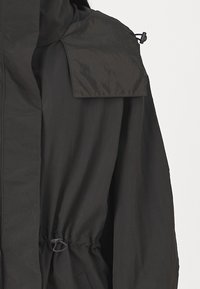 Veste imperméable noire avec un col haut, cordon de serrage ajustable à la taille et poche à rabat sur l'épaule. Texture lisse.