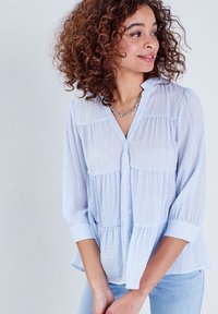 Blouse bleu clair, à boutons, avec des rayures verticales blanches, un design en étages et un tissu doux. Elle présente un col et des manches trois-quarts.
