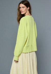 Prevelik pulover svetlo zelene barve z spuščenimi rameni in rebrastimi manšetami, kombiniran s plisirano svetlo rumeno krilo, pogled od zadaj.