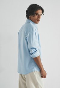 Gaiši zils krekls ar garām piedurknēm, kas sarullētas, izliekta apakšdaļa un mīksta auduma tekstūra. Šaura piegriezuma dizains, kuru valkā modelis, kas nedaudz pagriežies.