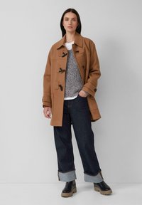 Frau steht vor einer hellgrauen Wand und trägt einen beige farbenen Toggle-Mantel, einen grauen Strickpullover, dunkle, aufgekrempelte Jeans und schwarze Stiefel mit beigen Sohlen.