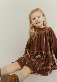 Brun fløjlskjole med samlede lag og lange ærmer. Har en glat tekstur og en høj halsudskæring, suppleret med knæhøje strømper og sko.