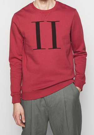 Homme portant un sweat-shirt rouge à manches longues avec un numéro romain II noir sur le devant et un pantalon ample gris, une main dans la poche.