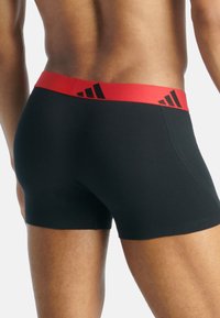 Černé boxerky s červeným elastickým pasem, na kterém jsou detaily černého loga Adidas. Hladká tkanina s přiléhavým střihem.
