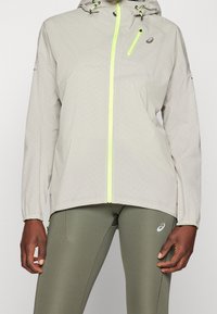 Persona que lleva una chaqueta con capucha de un beige claro con cremallera amarilla neón y leggings deportivos verde oliva con logo en el muslo.
