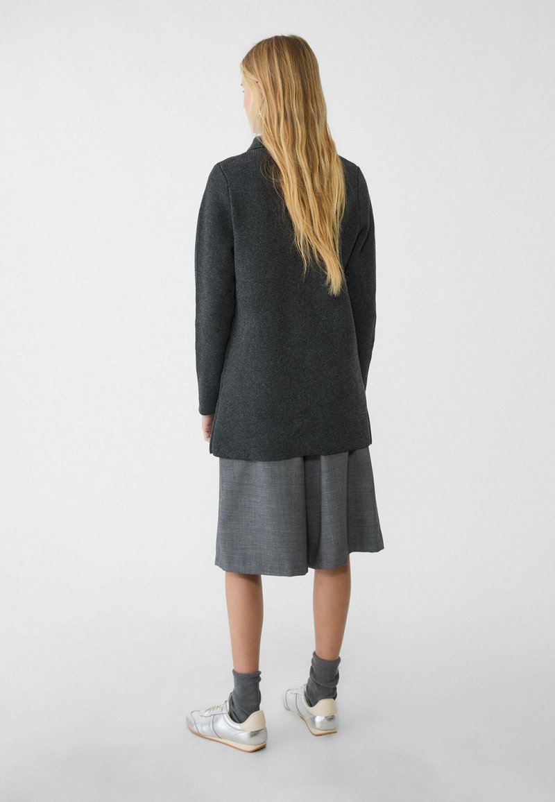 Un pull gris charbon à manches longues avec un ourlet légèrement courbé, porté sur une jupe grise longue jusqu'au genou, associé à des baskets argentées et des chaussettes grises.