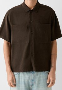 Mężczyzna ubrany w ciemny brązowy koszulowy t-shirt z krótkim rękawem, posiadający dwie kieszenie na piersi, oraz jasnoniebieskie dżinsy.