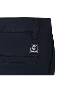 Pantaloni di tessuto blu navy con una texture liscia, dotati di una patch con il logo Timberland vicino alla vita e un dettaglio sulla tasca posteriore.