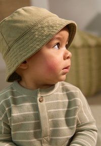 Cappello a secchiello color sabbia, camicia verde medio strutturata con righe bianche e bottone. Profilo di un bambino con viso rotondo e occhi grandi. Sfondo bianco.