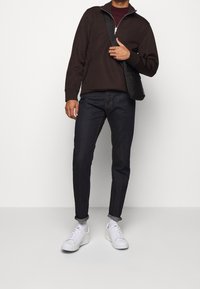 Man med mörkbrun zip-up tröja, mörka jeans med uppvikta ben, vita sneakers och svart axelremsväska framför en helt vit bakgrund.