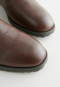 Botas de cuero marrón con puntera redondeada, superficie texturizada y suela de goma negra contrastante con un detalle estriado alrededor del borde.
