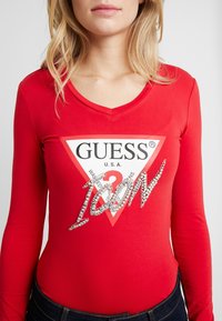 Bodysuit rojo de manga larga con escote en V, parche de logo triangular y texto "ICON" adornado con piedras brillantes. Tela elástica, estilo ajustado.