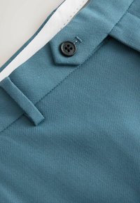 Gros plan sur un pantalon en tissu bleu sarcelle montrant la ceinture avec un bouton noir, une passants de ceinture et des coutures de doublure intérieure blanche.