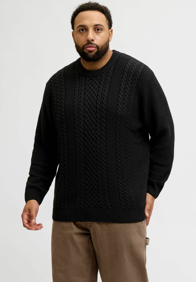 Black knit sweater featuring textured cable patterns, round neckline, and ribbed cuffs and hem. Worn with beige trousers.