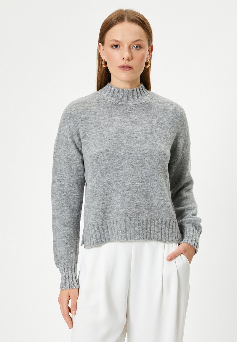 Grau gestrickter Pullover mit einem gerippten hohen Kragen und Bündchen, der eine kastige Passform und eine dezente Textur aufweist, kombiniert mit hellen Hosen.