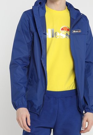 Person iført en blå lynlåshættetrøje over en gul ellesse-logo t-shirt og matchende blå bukser.