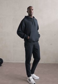 Black hoodie with a front pocket, relaxed fit; paired with matching black sweatpants. Model wears white athletic shoes. Simple design.