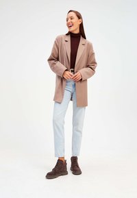 Blazer surdimensionné marron avec des revers, associé à un turtleneck marron ajusté, un jean droit bleu clair et des bottes en daim marron foncé à la cheville.
