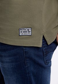 Polo verde oliva in tessuto testurizzato, con un'etichetta ricamata di colore blu navy con la scritta "JACK'S SPORTSWEAR" sull'orlo, abbinato a jeans blu scuro.