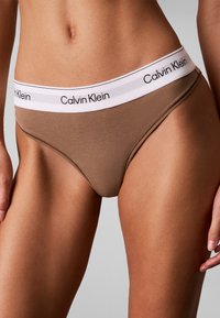 Γυναίκα που φορά καφέ εσώρουχα Calvin Klein με άσπρη ζώνη μέσης που έχει το όνομα της μάρκας με μαύρο κείμενο.