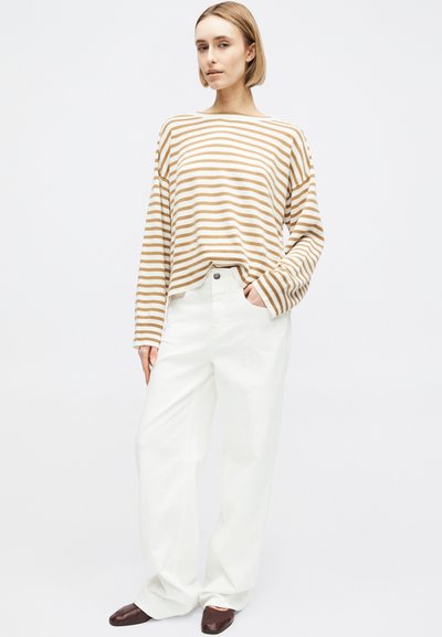 Femme debout, main dans la poche, portant une chemise à manches longues beige à rayures blanches, un pantalon blanc ample et des chaussures marron foncé, sur fond blanc.