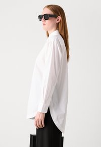 Camicia bianca a maniche lunghe con orlo curvo, polsini con bottone e colletto strutturato, abbinata a occhiali da sole neri e pantaloni neri.