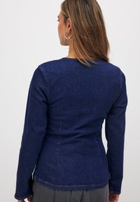 Tumši zilā džinsa jaka ar garām piedurknēm, piegulošu formu, redzamām šuvēm un noapaļotu apkakli. Tai ir gluda tekstūra.