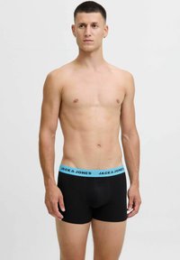 Moški stoji v črnih boksericah s svetlo modrim pasom, označenim s "JACK & JONES", na enobarvni beli podlagi.