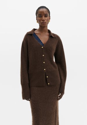 Femme portant un cardigan marron duveteux boutonné et une jupe assortie avec un haut en dentelle bleu marine visible en dessous, debout devant un fond uni.