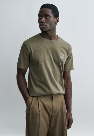 Bărbat cu păr scurt și creț, purtând un tricou verde măsliniu și pantaloni maro largi, stând cu mâinile în buzunare, uitându-se în lateral.