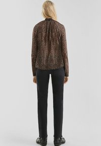Vrouw gezien vanuit de achterkant, gekleed in een bruin bloemetjesblouse met lange mouwen, zwarte rechte jeans en zwarte enkellaarsjes, tegen een effen achtergrond.