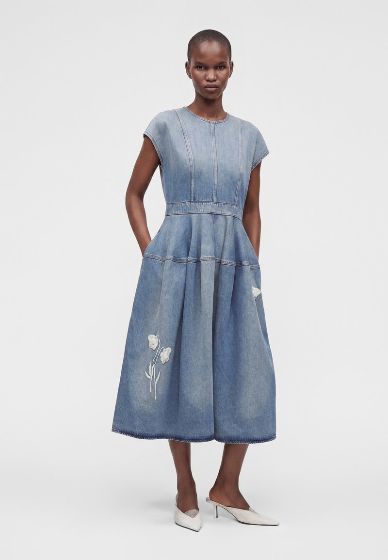 Donna che indossa un vestito midi in denim blu con maniche a cappuccio, ricamo floreale e tasche, abbinato a scarpe bianche con tacco alto e punta affilata.