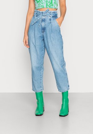 Persona que lleva pantalones de mezclilla azul claro de talle alto con cinturón anudado, combinados con botines verdes brillantes y un top corto verde con estampado.